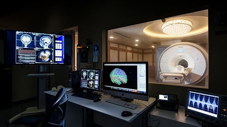 An MRI scanner behind a window, with brain images on the analysis workstation screens